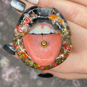 May include: A resin art piece shaped like a pair of lips with a tongue sticking out. The lips are black with gold flakes and colorful accents. The tongue is pink with a gold barbell piercing.