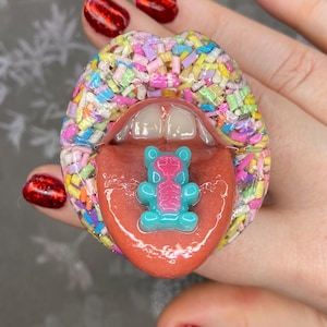 May include: A whimsical, colorful lip-shaped sculpture with a tongue and teeth. The lips are covered in a sprinkle-like design. A blue and pink gummy bear is on the tongue.