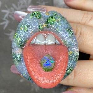 May include: A unique, artistic sculpture of lips with a red tongue and a gemstone. The lips are a translucent blue with green and gold accents. The gemstone on the tongue has blue and green hues.