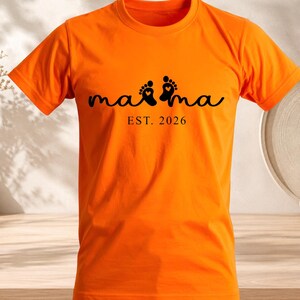 Peut inclure: T-shirt orange vif avec le mot "mama" en écriture noire, avec deux empreintes de pieds avec des détails de cœur. En dessous du mot "mama" se trouve le texte "EST. 2026". Le t-shirt est en matière souple.