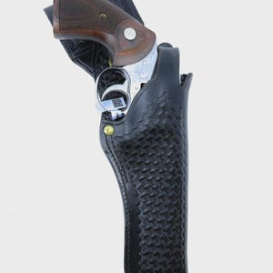 Peut inclure: Un &eacute;tui en cuir noir avec un motif tiss&eacute; contient un revolver argent&eacute; avec une poign&eacute;e en bois marron. L'&eacute;tui a une fermeture &agrave; bouton-pression et des rivets en laiton. Le revolver est partiellement visible.