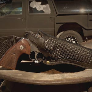 May include: A revolver with a brown wooden grip and a silver barrel rests in a black leather holster with a woven pattern. The holster is on a wooden surface, with a vehicle in the background.