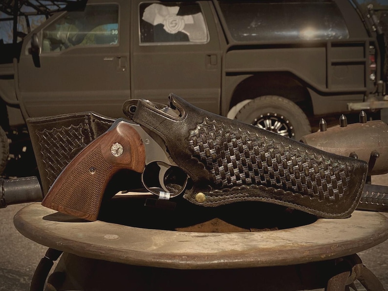 Peut inclure: Un revolver avec une poign&eacute;e en bois marron et un canon argent&eacute; est pos&eacute; dans un &eacute;tui en cuir noir avec un motif tiss&eacute;. L'&eacute;tui est sur une surface en bois, avec un v&eacute;hicule en arri&egrave;re-plan.