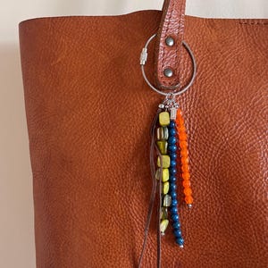 Könnte beinhalten: Eine braune Ledertasche mit einem dekorativen Perlenanhänger. Der Anhänger hat Stränge aus blauen, orangefarbenen und gelben Perlen sowie kleine Lederquasten. Die Tasche hat eine strukturierte Oberfläche und einen Ledergriff.