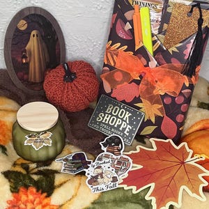 May include: A gift set featuring a wrapped book with an orange ribbon, a green candle with a wooden lid, a small crocheted pumpkin, a ghost-themed oval plaque, a leaf-shaped cutout, and several stickers including one reading Book Shoppe.