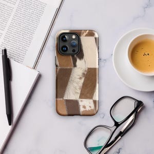 Peut inclure: Coque de smartphone avec un motif patchwork dans les tons marron, beige et blanc. L'étui présente un aspect texturé, avec un mélange de sections lisses et poilues. L'étui est posé sur une surface en marbre.