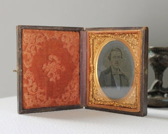 May include: A vintage, wooden, hinged photo frame with a velvet interior and a gold-toned frame surrounding a black and white portrait of a man. The frame is open and the portrait is visible.