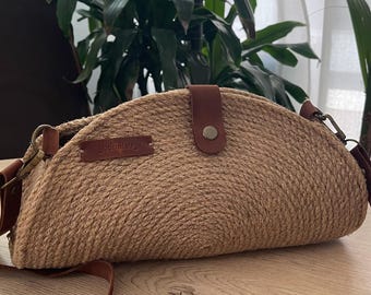 May include: A semi-circular handbag crafted from coiled tan jute rope, featuring a brown leather strap and a matching leather closure tab with a metal snap button. A small rectangular brown leather tag embossed with the text boholab is attached.
