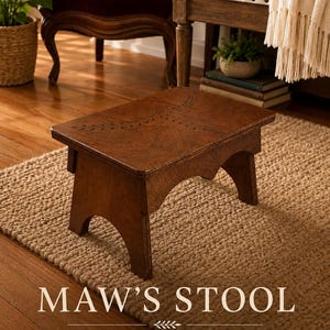 May include: A dark brown wooden step stool featuring a rectangular top with a carved leaf pattern and arched side supports. The text MAW'S STOOL is printed in white at the bottom, followed by the text SOLID. STURDY. BEAUTIFULLY CRAFTED. THE PERFECT LITTLE STEP FOR EVERY HOME.