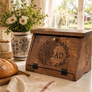 May include: A dark brown wooden bread box with a hinged front door featuring a brass knob, black metal hinges, and an engraved design of a wreath, a heart, and the word BREAD. A small goose illustration is etched on the right side panel.