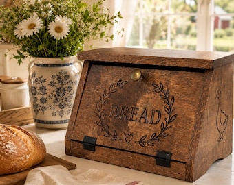 Könnte beinhalten: Ein dunkelbrauner Brotkasten aus Holz mit einer aufklappbaren Fronttür, einem Messingknopf, schwarzen Metallscharnieren und einer Gravur, die einen Kranz, ein Herz und das Wort BREAD zeigt. Auf der rechten Seite ist eine Gans eingraviert.
