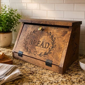 May include: A dark brown wooden bread box with a slanted front door featuring a brass knob, black metal hinges, and an engraved laurel wreath design surrounding the word BREAD and a small heart symbol centered on the front panel.