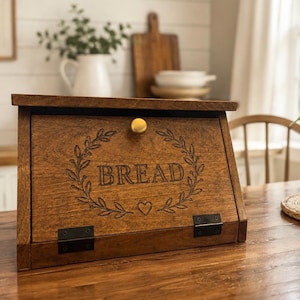 May include: A medium-brown wooden bread box with a slanted front door featuring a gold knob. The door is engraved with the word BREAD centered inside a leafy wreath design with a small heart below. Two black metal hinges secure the door base.