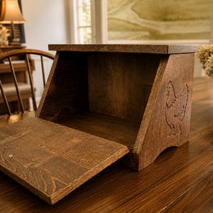 May include: A dark brown wooden bread box with a hinged front door that folds downward. The side panel features a laser-engraved outline of a chicken. The box has a rectangular top, a flat base, and a smooth, stained wood grain finish throughout.