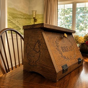 May include: A dark brown wooden bread box with a slanted front door featuring a brass knob. The front panel is engraved with a wreath, a small heart, and the word BREAD. The side panel displays an engraved chicken silhouette. Black metal hinges.