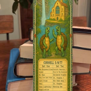May include: A decorative concert poster featuring two cartoon turtles in front of a waterfall and a yellow building. The poster lists the setlist for a Cornell concert on 5/8/77. The poster is framed with an ornate green border.