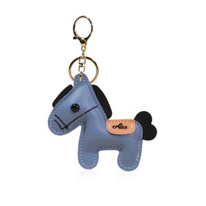 May include: A blue horse-shaped keychain with black mane and tail, and a gold-colored keyring. The keychain has the name "Alice" on a tan saddle. The horse is made of stitched leather-like material.
