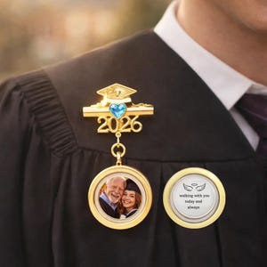 May include: A gold graduation pin with a blue heart-shaped gem and the year "2026". The pin has a small graduation cap design. A round pendant with a photo of a smiling couple hangs below. Another round pendant says "walking with you today and always".