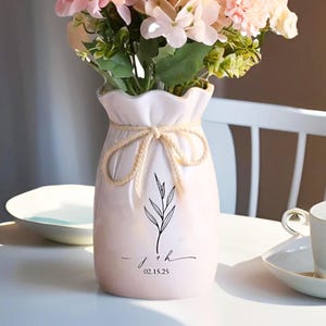 Puede incluir: Un jarrón de cerámica blanca con un detalle de cuerda anudada y un diseño de hojas negras. El jarrón contiene flores artificiales rosas y verdes. La parte inferior del jarrón tiene las iniciales "j + h" y la fecha "02.15.25".