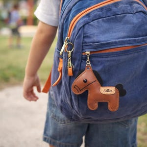 May include: A brown leather horse keychain with the name "Alice" on a small tag, attached to a blue backpack with orange accents. The keychain has black details and a gold-colored metal clasp.