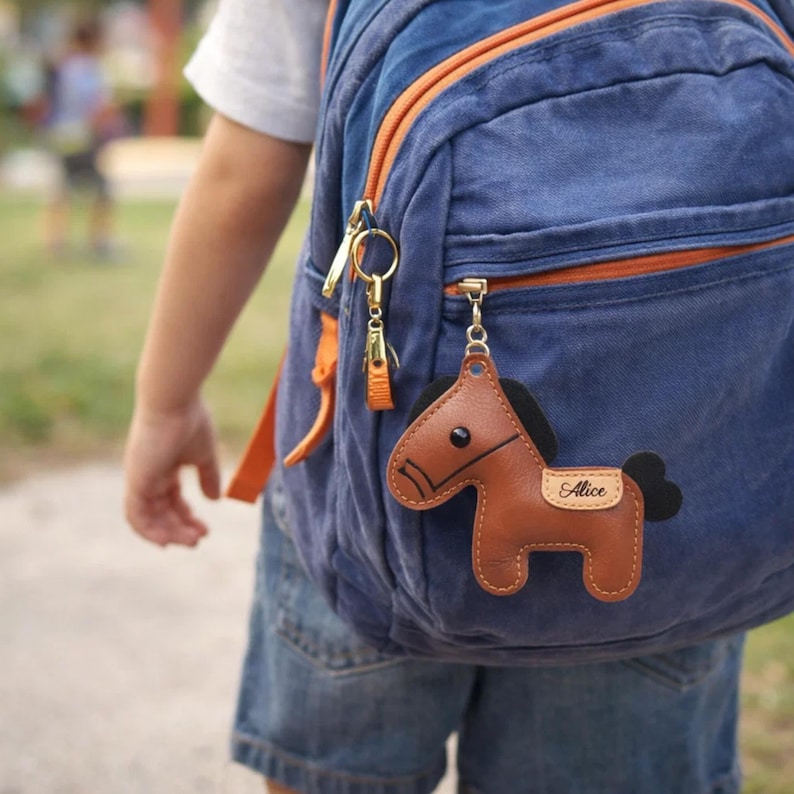 May include: A brown leather horse keychain with the name "Alice" on a small tag, attached to a blue backpack with orange accents. The keychain has black details and a gold-colored metal clasp.