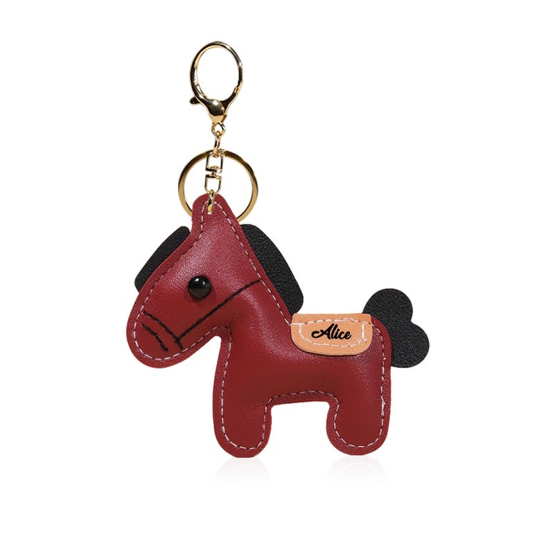 May include: A burgundy leather horse keychain with a gold-colored clasp. The horse has black accents for the mane and tail, and the name "Alice" is printed on a tan patch. The keychain is designed with white stitching.