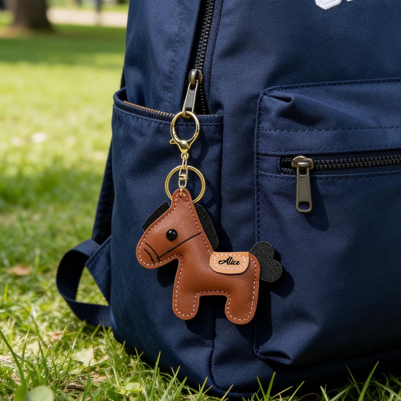 May include: A brown leather horse keychain with black mane and tail, attached to a gold-colored keyring. The keychain is personalized with the name "Alice" on a small tag. The keychain hangs from a navy blue backpack.