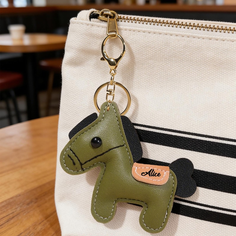 May include: A green leather horse keychain with black mane and tail, and a small tan saddle with the name "Alice" on it. The keychain has a gold-colored clasp and ring, attached to a cream-colored bag with black stripes.