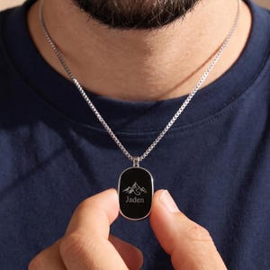 May include: A silver-toned chain necklace featuring an oval-shaped pendant with a black face. The pendant is engraved with a white line-art illustration of mountain peaks above the name Jaden in a clean, serif font centered on the surface.