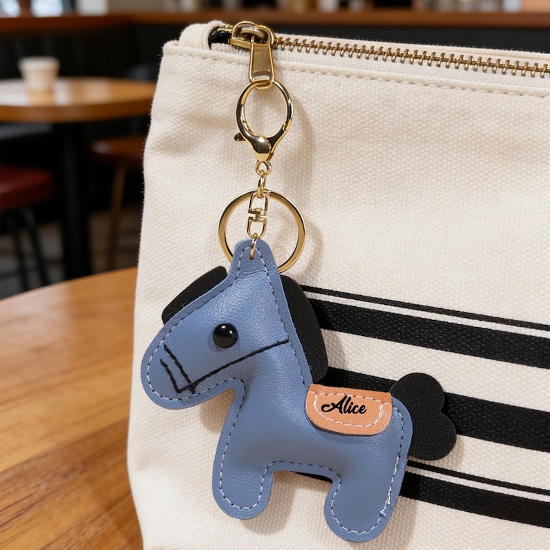 May include: A blue leather horse keychain with black mane and tail, and a tan saddle with the name "Alice" printed on it. The keychain has a gold-colored clasp and ring, attached to a cream-colored bag with black stripes.