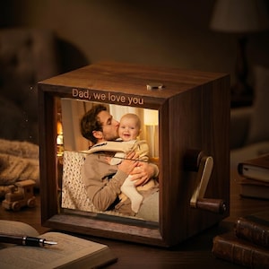 Könnte beinhalten: Eine quadratische Spieluhr aus Holz mit dunkelbraunem Finish und einer Handkurbel an der Seite. Die Vorderseite zeigt ein Foto eines Erwachsenen, der ein Baby hält, mit dem eingravierten Text "Dad, we love you" am oberen Rand des Holzrahmens.
