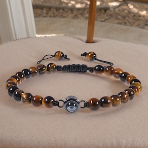 May include: A bracelet made of tiger's eye and black beads, with a central silver-coloured charm with a blue eye design. The adjustable cord allows for a comfortable fit. The bracelet is displayed on a neutral-toned surface.