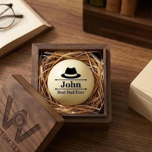 Puede incluir: Un juego de regalo personalizado con una esfera crema con un sombrero negro, el nombre "John" y el texto "Best Dad Ever". La esfera descansa en una caja de madera llena de paja. La caja está parcialmente abierta, revelando una tapa de madera.
