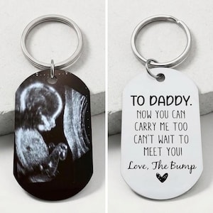 May include: A silver keyring with two oval pendants. One shows a grayscale ultrasound image. The other pendant has the text "TO DADDY. NOW YOU CAN CARRY ME TOO CAN'T WAIT TO MEET YOU! Love, The Bump" with a heart.
