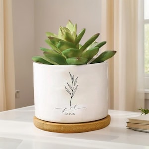 Op de afbeelding: Een witte keramische plantenpot met een groene vetplant. De pot heeft een zwart bloemmotief en de inscriptie "j + h 02.15.25". Hij staat op een ronde, natuurlijke houten onderzetter. Woondecoratie.