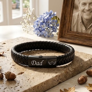 Può includere: Un bracciale in pelle intrecciata nera con una chiusura rettangolare in metallo. La chiusura è incisa con la parola "Dad" in carattere corsivo e un piccolo grafico di ali d'angelo con un'aureola sopra, centrato sulla parte anteriore in metallo.
