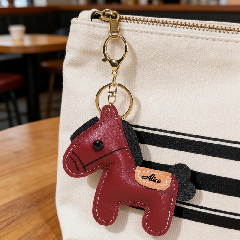 May include: A burgundy leather horse keychain with black mane and tail, and the name "Alice" on a tan patch. The keychain has a gold-colored ring and clasp, attached to a cream-colored bag with black stripes.