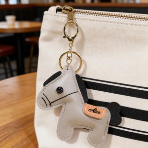 May include: A gray leather horse keychain with black mane and tail, attached to a gold-colored clasp and ring. The keychain is hanging in front of a cream-colored canvas bag with black stripes. The word "Alice" is on the horse.