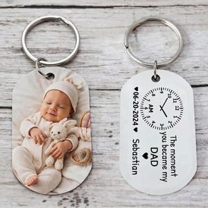 May include: Two metal keychains. One features a baby photo, the other has a clock design with the text "The moment you became my DAD" and the date "06-20-2024". Both keychains have silver rings.