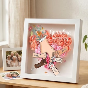 May include: A white framed shadow box with a floral arrangement and the words "Grandma WE LOVE YOU". The box features hands and paws holding each other, with names like "Bella" and "Buddy".