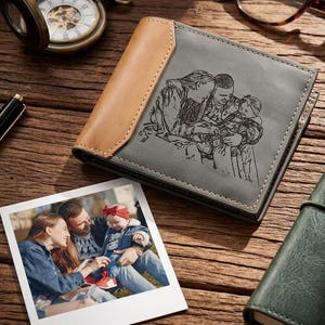 Puede incluir: Cartera de cuero gris con un grabado de retrato familiar y un acento de cuero marrón claro. La cartera es cuadrada y tiene un borde cosido. Una foto de una familia está al lado de la cartera.