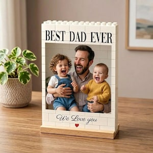 以下が含まれることがあります： 上部に「BEST DAD EVER」、下部に小さな赤いハートと共に「We Love you」の文字が記された、白いブロック製の長方形フレーム。木製の台座の上に、男性が2人の幼い子供を抱いている写真が収められています。