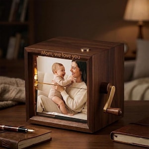 May include: A wooden music box with a photo of a mother and child. The box has a crank and a small brass-colored knob on top. The text "Mom, we love you" is engraved on the top. The box is sitting on a table with a pen and a notebook.