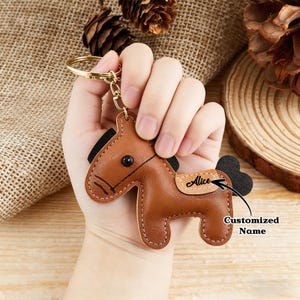 May include: A brown leather horse keychain with a gold-colored keyring. The horse has black accents for the mane and tail, and a stitched design. The name "Alice" is printed on the horse's side. The words "Customized Name" are also visible.