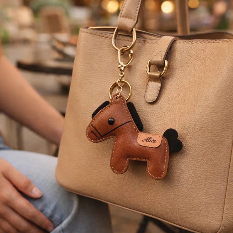 May include: A brown leather horse keychain with black mane and tail, attached to a gold-colored keyring. The keychain is personalized with the name "Alice" on a small tag. The keychain hangs from a beige handbag.