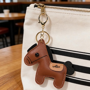 May include: A brown leather horse keychain with a gold-colored clasp and ring. The horse has a black mane and tail, and the name "Alice" is printed on a small tan patch. The keychain is attached to a white canvas bag with black stripes.