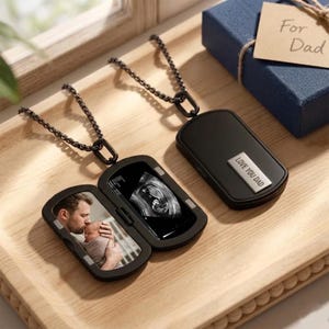 Op de afbeelding: Zwarte dog tag hanger ketting met een scharnierend compartiment. Eén hanger toont een foto van een vader en baby, de andere een echo-afbeelding. De gesloten hanger heeft "LOVE YOU DAD" op een zilveren plaat.