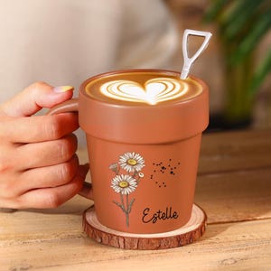 Puede incluir: Una taza de color terracota con forma de maceta, llena de un café con leche con un diseño en forma de corazón. La taza tiene un asa y una cuchara pequeña. Está decorada con ilustraciones de margaritas y el nombre "Estelle". La taza descansa sobre un posavasos de madera.