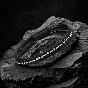 Könnte beinhalten: Schwarzes Lederarmband mit silberfarbenen Perlen und einem Magnetverschluss. Das Armband wird auf einer dunklen, strukturierten Oberfläche präsentiert, die sein modernes Design hervorhebt. Dieses Accessoire ist für den Alltag geeignet.