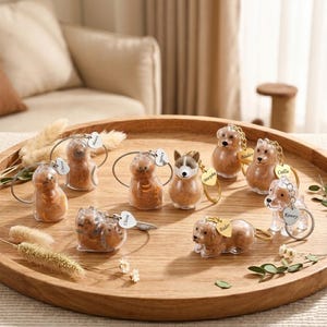 May include: A collection of clear resin keychains shaped like cats and dogs, each with a small gold-colored tag. The keychains are displayed on a wooden tray, with some featuring personalized names.
